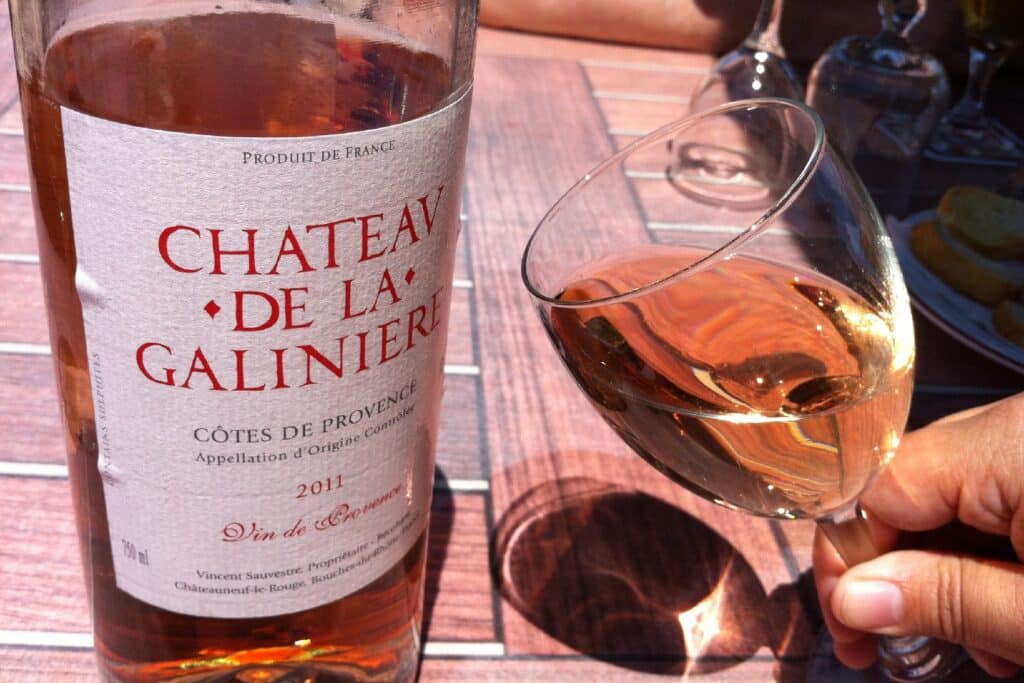 A hand holding a glass of rosé wine with a bottle of 'Chateau de la Galinière, Côtes de Provence 2011' in the background, reflecting a sunny outdoor setting, perfect for those seeking a taste of Marseille souvenirs.