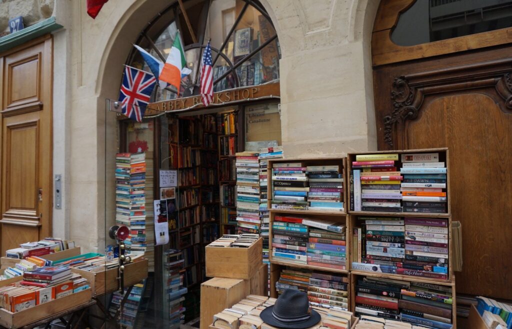 English Bookshops in Paris, A cozy and inviting second-hand bookstore, the Abbey Bookshop on a street in Paris, with a collection of books piled and shelved outside the shop. The shopfront is adorned with the British, Irish, and European Union flags above the entrance, which has a classic wooden door frame with intricate carvings. A hat is casually placed on one of the outdoor bookshelves, adding to the quaint charm of the scene.