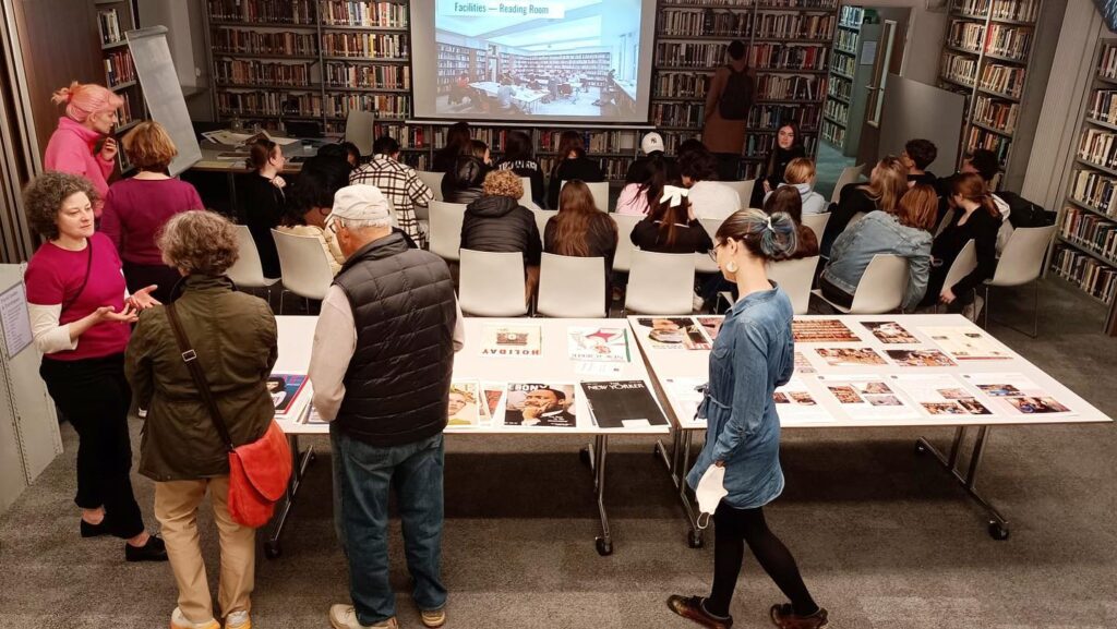 A busy event at the American Library in Paris, with attendees engaging in various activities. In the foreground, a woman in a denim shirt is observing posters laid out on a table, while behind her, a man in a vest converses with a woman in a green jacket. In the center, a group is seated facing a presentation screen, attentively watching. The room is lined with bookshelves filled with a vast array of books, creating an intellectual and communal atmosphere.