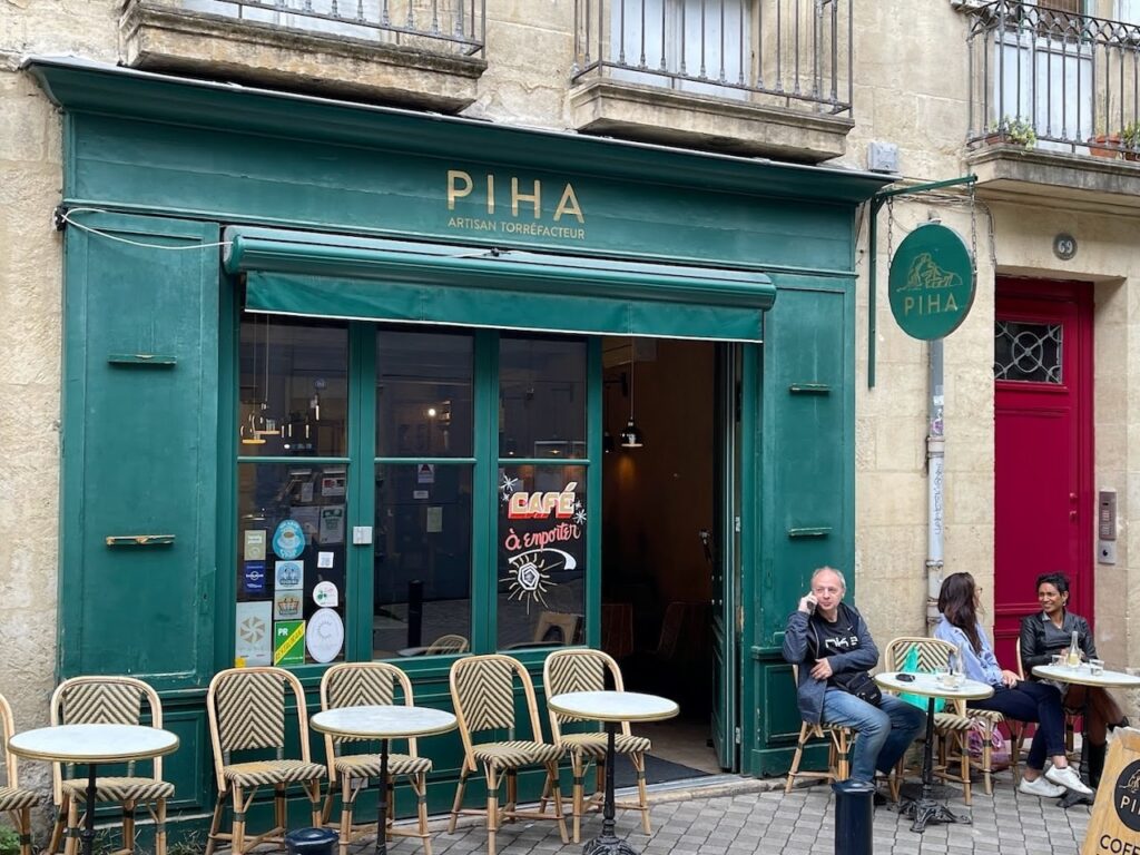 Customers enjoying their beverages at Café Piha, an artisan coffee roaster in Bordeaux, with a classic Parisian sidewalk seating arrangement. The café's green façade and logo are prominent, contributing to the relaxed, urban atmosphere perfect for a day in Bordeaux.