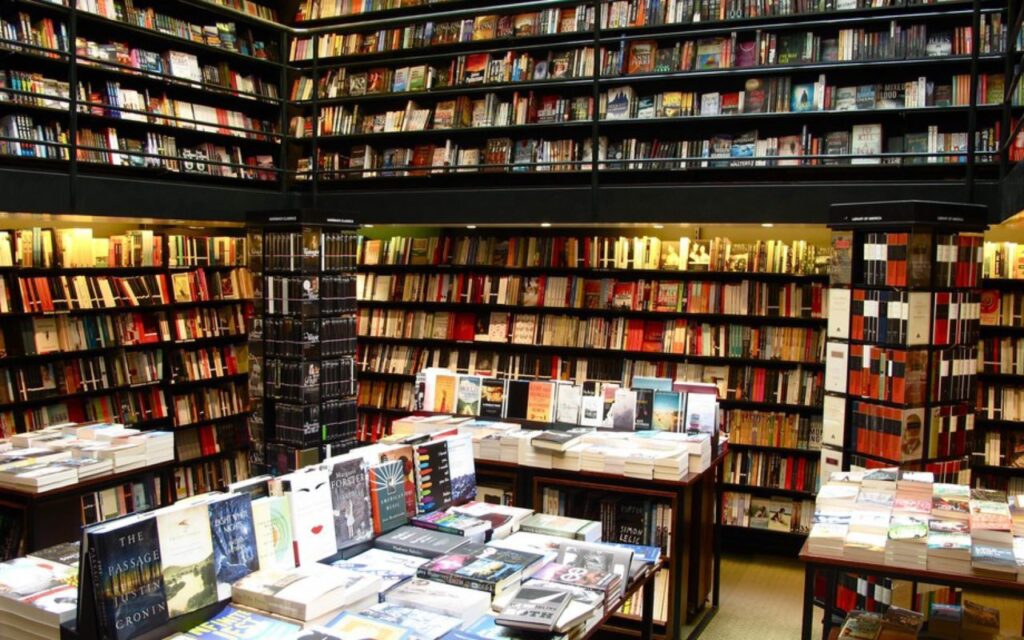 Interior of Librairie Galignani in Paris, showcasing its elegant and extensive book collection. The shelves are neatly organized and filled with books of various genres, featuring a sophisticated black shelving system that enhances the rich colors of the book spines. The warm lighting casts a soft glow over the space, highlighting the rows upon rows of literary works, while a central column wrapped with bookshelves adds a unique architectural element to the store's design.