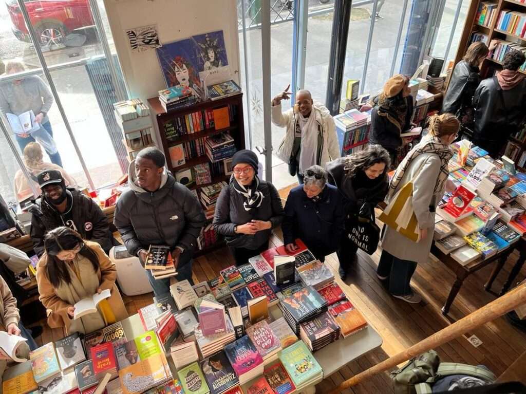 English Bookshops in Paris, A vibrant and bustling scene inside The Red Wheelbarrow bookstore in Paris. Diverse groups of people, including a man cheerfully waving, are browsing and selecting books from the abundant collection displayed on wooden tables and shelves. The interior is well-lit with natural light streaming in from the large windows, reflecting the lively atmosphere of this literary hub.