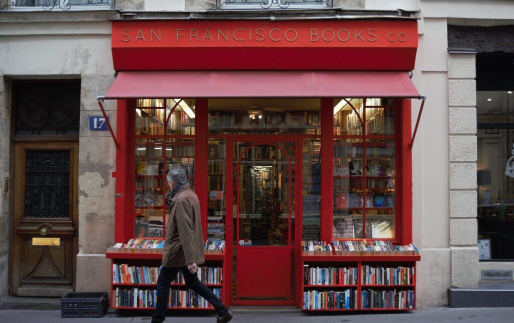 The storefront of San Francisco Book Company in Paris, France, featuring a vivid red facade and awning. A man in a brown jacket is walking past the shop, where bookshelves full of books are displayed both inside the window and outside along the street. The warm lighting from within and the inviting open door suggest a welcoming atmosphere for book lovers and passersby.
