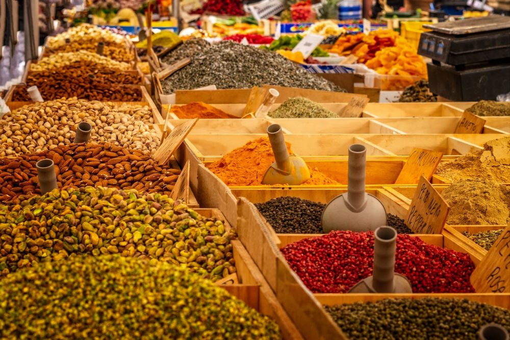 Vibrant display of spices and nuts at a Marseille market, with heaps of almonds, pistachios, and an array of colorful spices in wooden crates, invoking the senses and ideal as Marseille souvenirs.