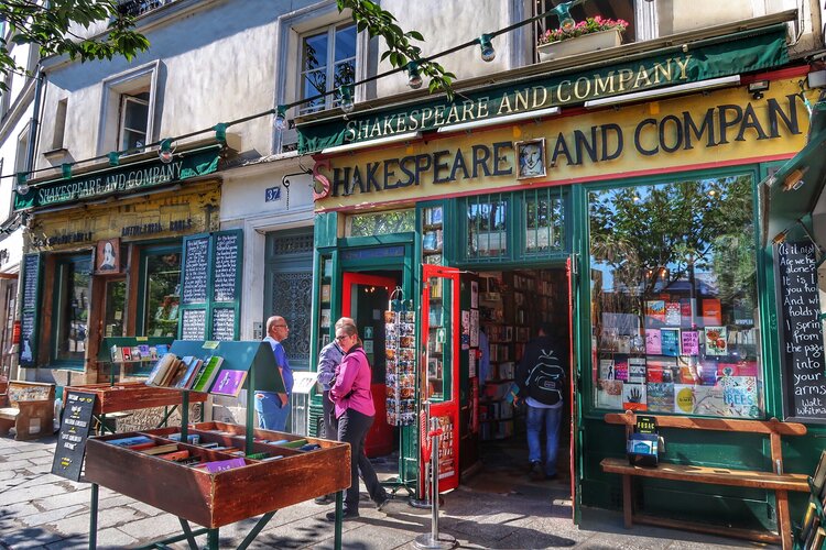 English Bookshops in Paris with The iconic Shakespeare and Company bookstore in Paris, France, with its distinctive green and yellow facade and colorful signage. A gentleman in a purple shirt is walking by while another person browses inside. The bookstore's front display features a variety of books, and a vintage red phone booth adds to the quaint, literary charm of the scene. Green foliage drapes above the entrance, inviting visitors into this haven of literature.
