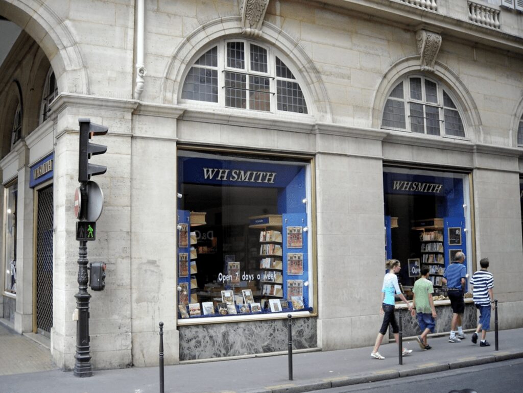 English Bookshops in Paris, The front of W.H. Smith bookstore in Paris, France, set in an elegant stone building with arched windows. The shop's deep blue awnings and signage contrast with the light stone facade. Pedestrians, including a family with children, are walking past the store, some glancing at the window displays featuring a selection of books. A traditional Parisian street lamp and a green pedestrian traffic light at the corner add urban details to the scene.