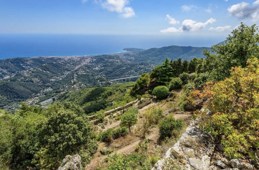 A panoramic view from a mountain path reveals a verdant landscape that descends to a coastal town, with the azure blue sea meeting the horizon under a bright sky, showcasing the natural beauty of the Mediterranean region.