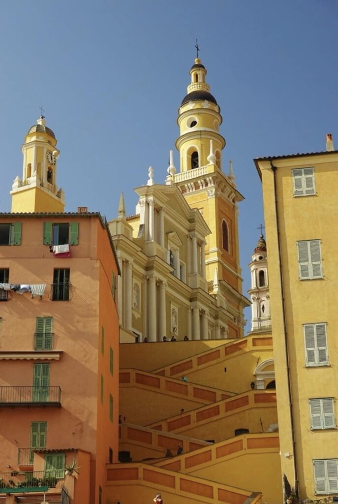 Things to do in Menton: The photo captures the striking architecture of a baroque-style church with ornate yellow and white towers reaching into the clear blue sky, juxtaposed against the warm tones of surrounding Mediterranean buildings and staircases.