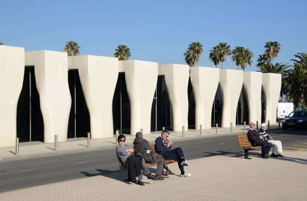 This image depicts a sunny, urban promenade where people relax on benches, with a unique modern building featuring wavy, abstract-shaped openings in the background, complemented by tall palm trees against a clear blue sky.