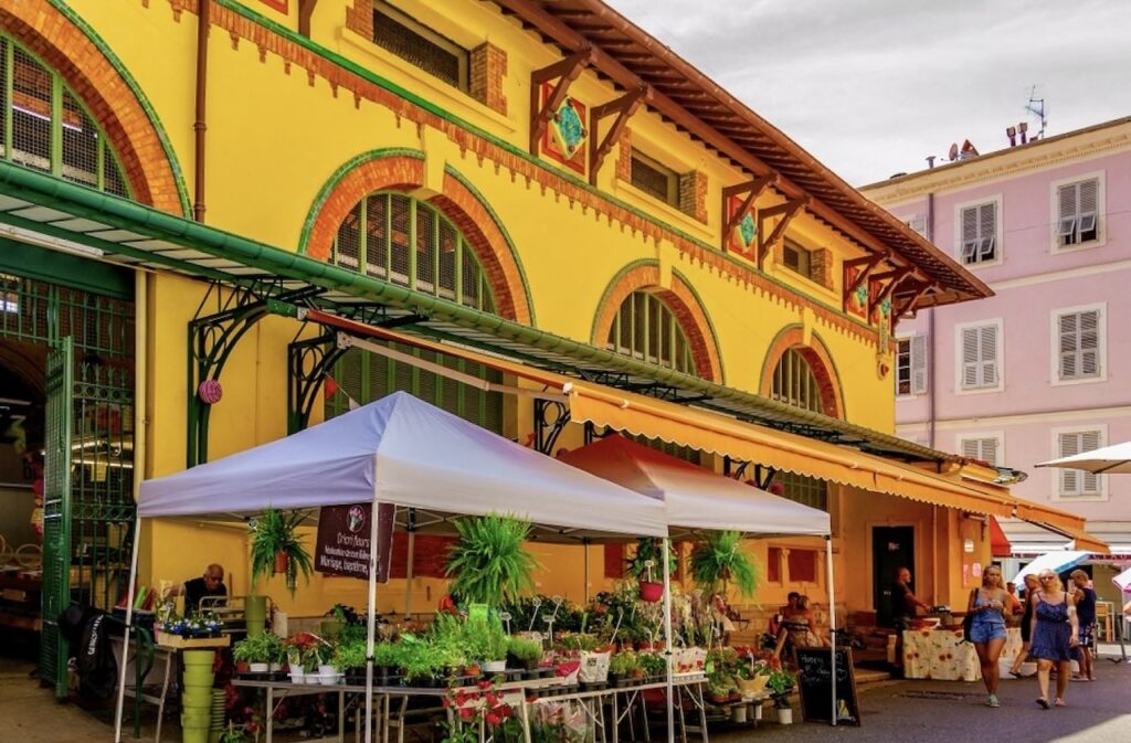 A bustling outdoor market under white canopies in front of the vibrant yellow Marché des Halles building in Menton, France, offering an array of plants and flowers, with locals and tourists strolling around, enjoying one of the popular things to do in Menton.