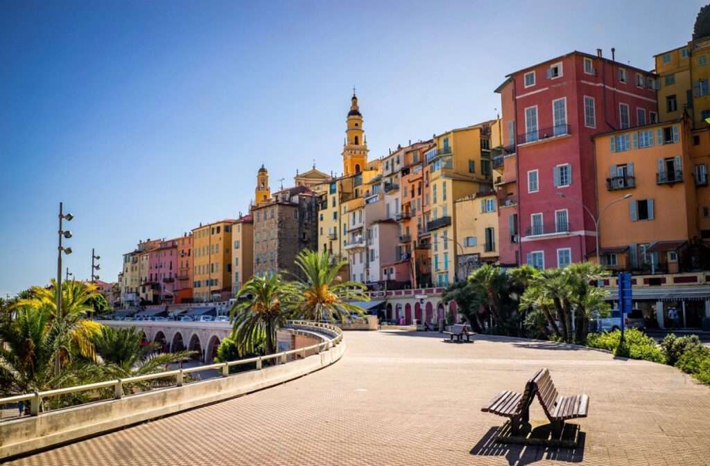 Things to do in Menton: The photo showcases a picturesque esplanade with a solitary bench, leading the eye towards a cluster of colorful buildings and a prominent church tower, all under the radiant Mediterranean sun, exemplifying the charm of a coastal European town.