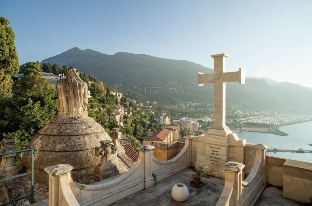 Things to do in Menton: This image shows a serene hilltop cemetery overlooking a coastal town with lush green mountains in the background. A prominent stone cross in the foreground symbolizes peace and eternity, with the Mediterranean Sea gently stretching into the horizon.