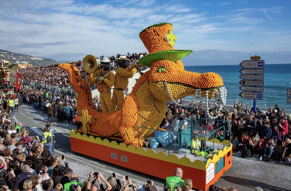 Things to do in Menton: A vibrant parade float featuring a large, orange dragon and musicians, all adorned with citrus fruits, entertains a crowd along the seaside promenade under a clear blue sky, capturing the festive spirit of a traditional fruit festival.