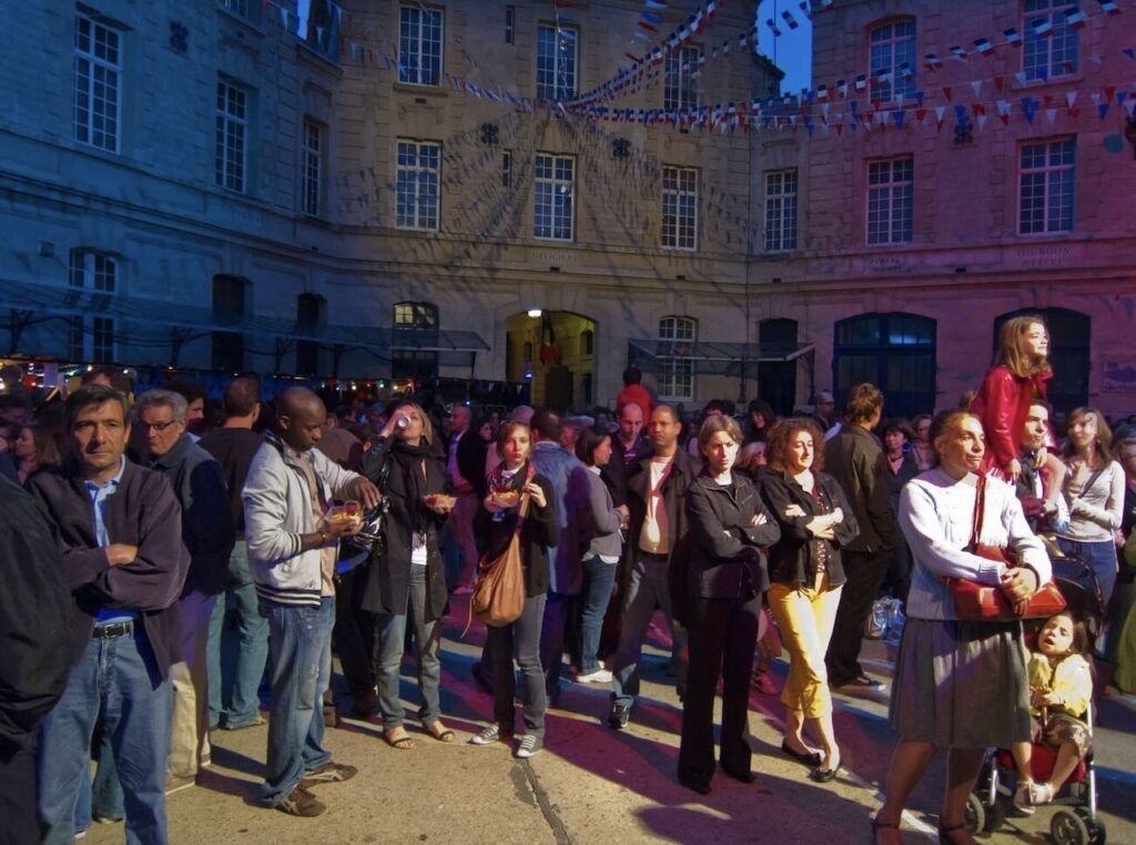 This image shows a diverse crowd of people gathered in a festive outdoor setting at dusk. They appear to be engaged in a social event, with some individuals holding drinks, suggesting a relaxed and convivial atmosphere. The background is adorned with stringed flags, indicating a celebration or public festivity, set against an illuminated historical building. The image captures a moment of community and shared enjoyment.