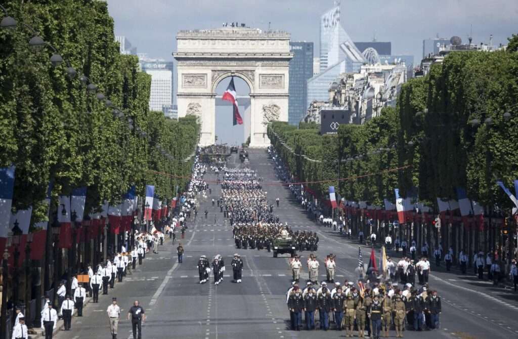 Bastille Day in Paris: This image shows a grand military parade taking place on a wide boulevard, which is the Champs-Élysées in Paris, with the Arc de Triomphe in the background. Military personnel are marching in formation, and vehicles are also part of the parade, indicating a formal and possibly national celebration or commemoration. The presence of the French flag and uniformed service members suggests a significant patriotic event. The scene is well-organized and ceremonial, reflecting a sense of national pride.