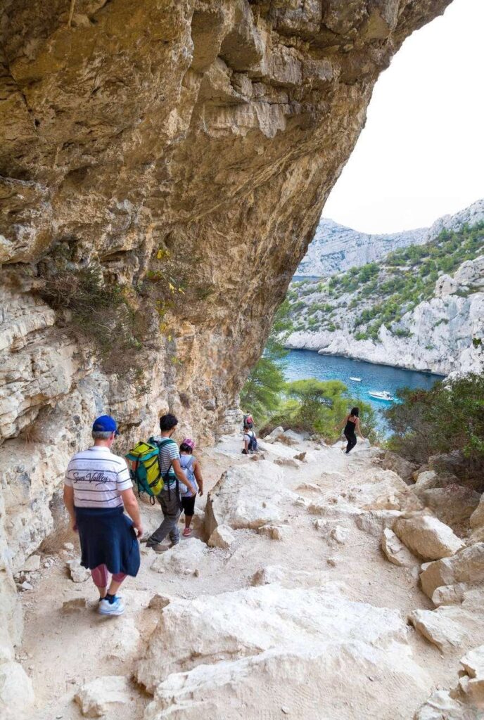 A group of hikers navigating a rugged trail in the Calanques National Park, with a scenic view of a turquoise cove and limestone cliffs in the distance. The foreground shows the rocky path and diverse vegetation, capturing the essence of outdoor adventure in Marseille.