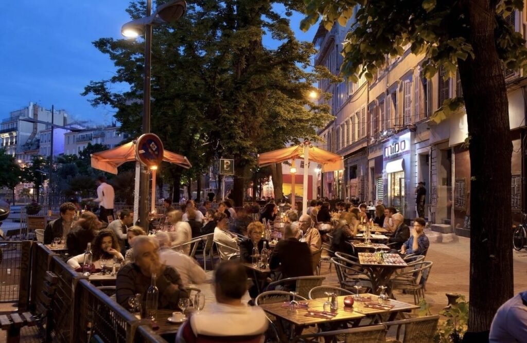 An evening at a bustling outdoor café in Marseille, with patrons engaged in conversation over dinner, warmly illuminated by overhead lights and street lamps, encapsulating the vibrant dining culture characteristic of One Day in Marseille.