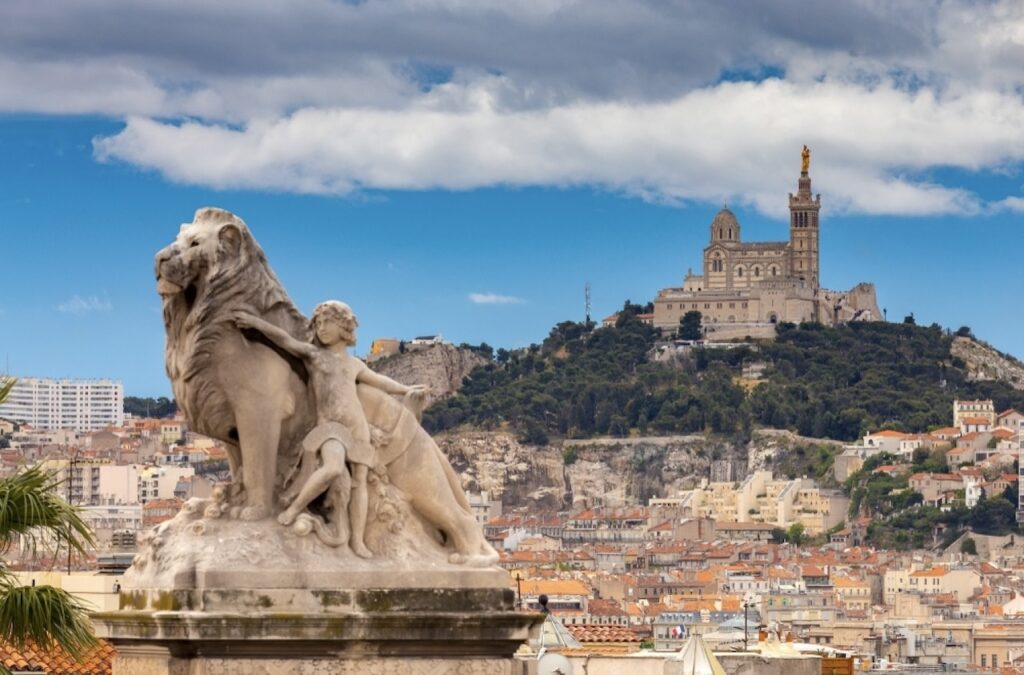 Sculpture of a lion and a child overlooking the cityscape of Marseille, with the historic Notre-Dame de la Garde basilica perched on a hill in the background, under a partly cloudy sky, perfect for exploring in one day in Marseille.