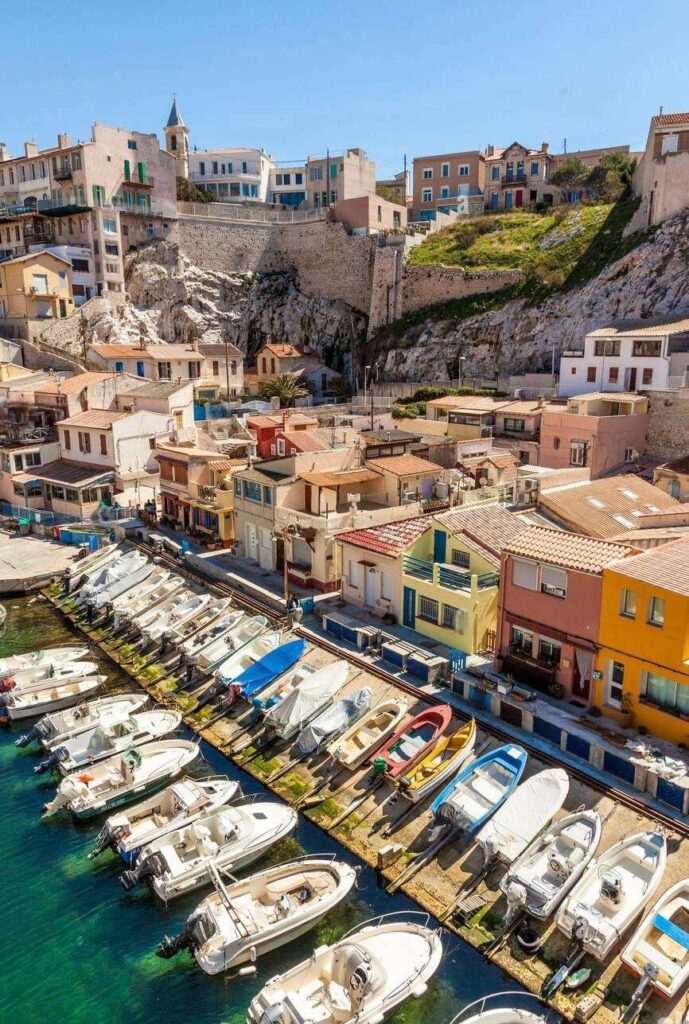 Vallon des Auffes, a picturesque fishing port in Marseille, with a row of small boats moored along the quay. The charming, colorful houses and the rugged cliffs with historical structures on top, beautifully depict this quaint Mediterranean seaside village.