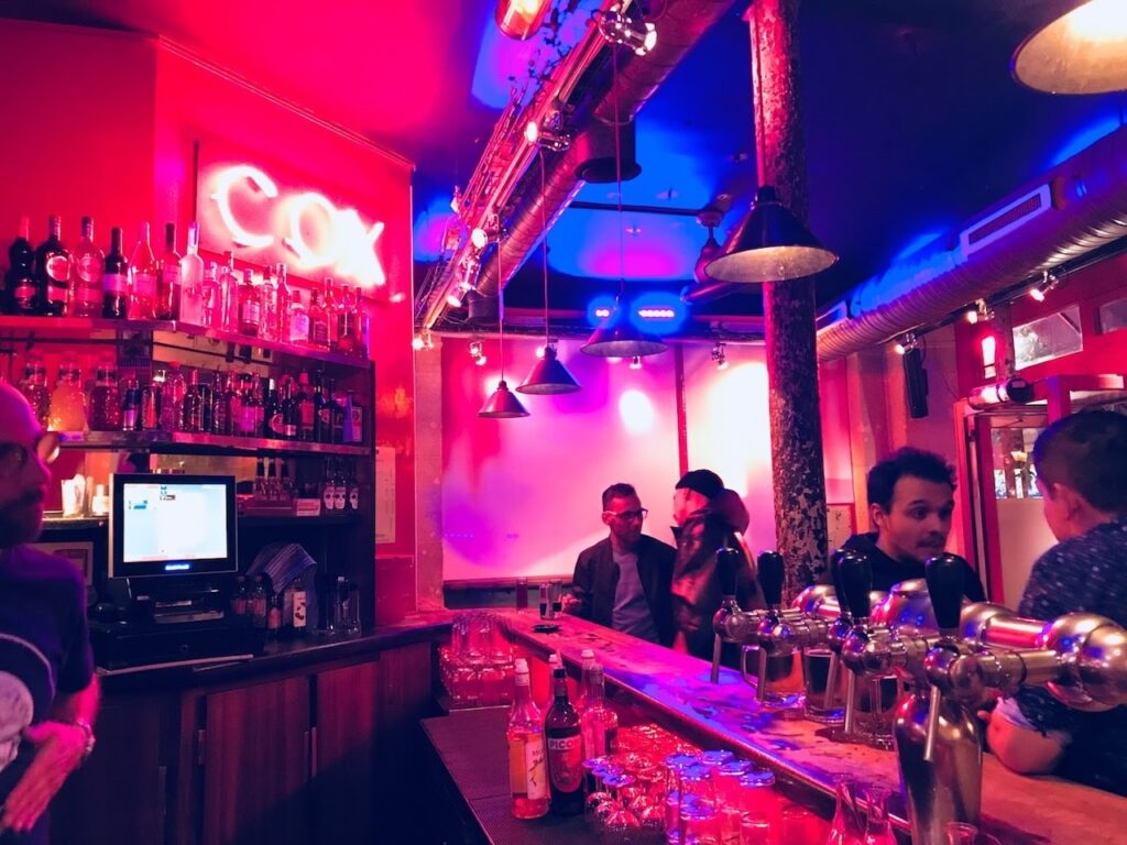 Patrons enjoy a lively evening at Cox bar in Paris, France, with its vibrant neon lighting and a well-stocked bar glowing under magenta hues, creating an energetic and inviting atmosphere.