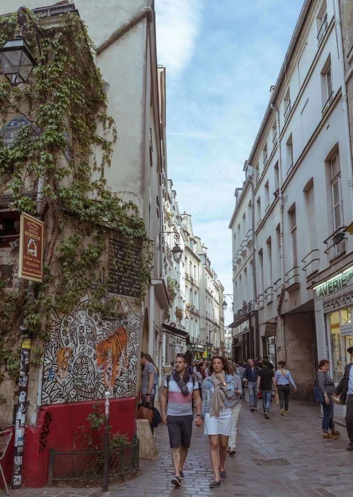 A typical day in the bustling Rue des Rosiers in Le Marais, Paris, with pedestrians strolling past ivy-covered buildings and striking street art, capturing the district's blend of historic charm and contemporary culture.