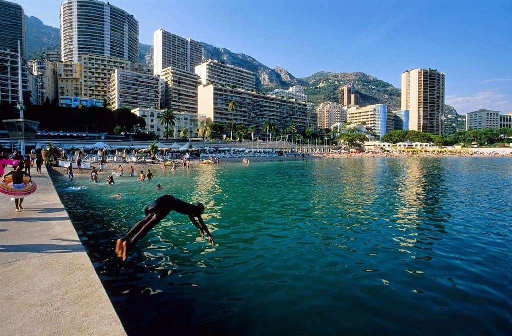 One Day in Monaco: A dynamic scene at Larvotto Beach with people swimming and lounging on the sand, while a person performs a dive into the crystal-clear Mediterranean Sea, all set against the backdrop of Monaco's urban skyline.