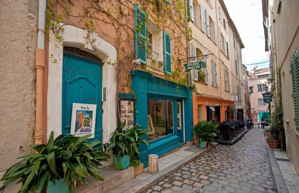 Quaint cobblestone street in Marseille with vibrant blue and teal storefronts, including a gallery with a 'Hors Série' poster, capturing the charming atmosphere ideal for Marseille souvenirs.