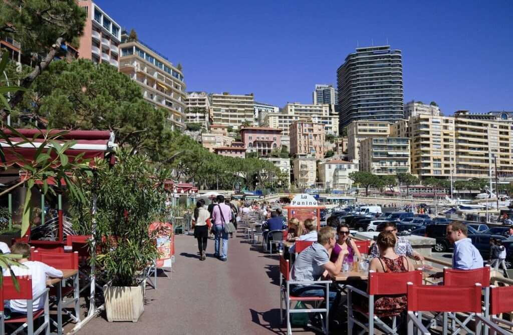 One Day in Monaco: Visitors and locals enjoy a sunny day along the promenade in Monte Carlo, with al fresco dining at waterside restaurants and a view of the bustling marina and Monaco's high-rise urban landscape.