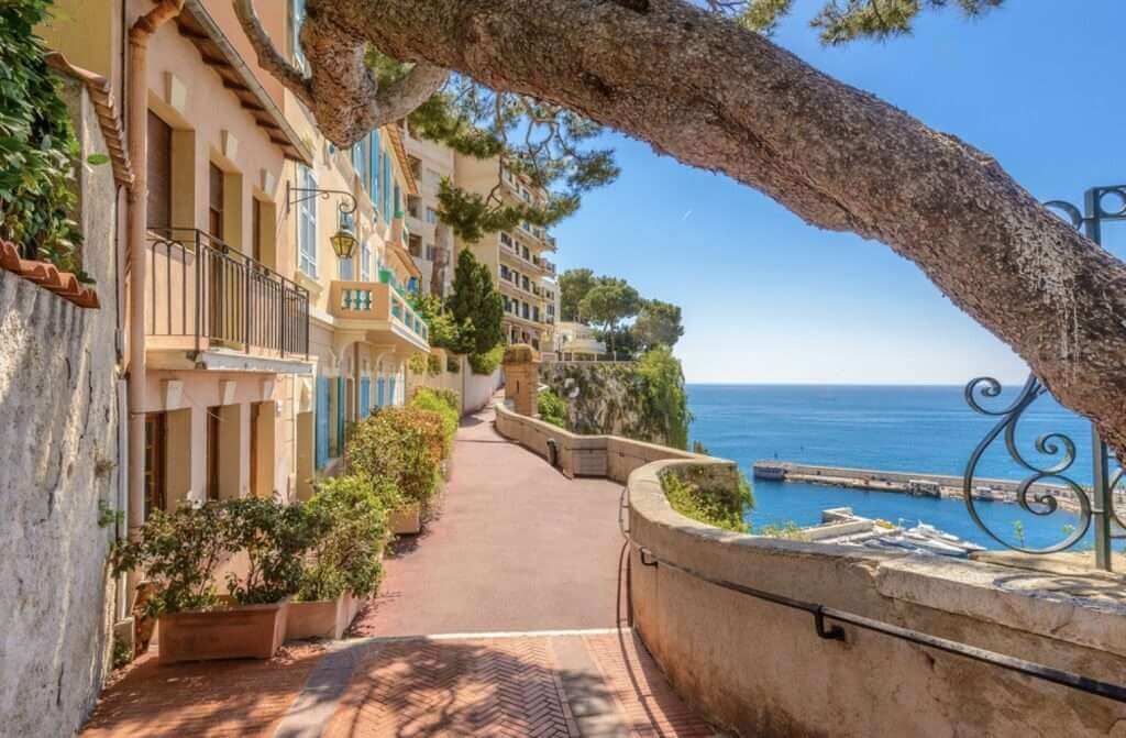 One Day in Monaco: A picturesque walkway in Monaco Village lined with traditional peach and yellow buildings, with the azure Mediterranean Sea peering through a natural archway formed by an overhanging tree, inviting exploration and discovery.