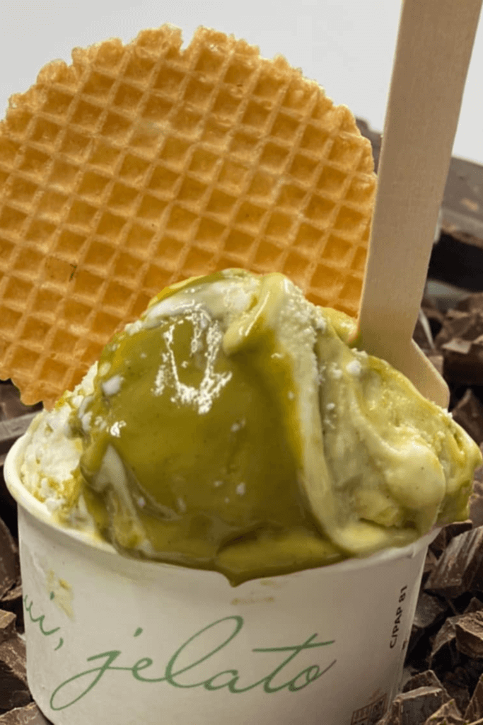 A paper cup with the best ice cream in Nice from Oui, Gelato, showcasing a luscious scoop of vibrant pistachio gelato, perfectly complemented by a crispy wafer and a wooden spoon, epitomizing the artisanal gelato experience.