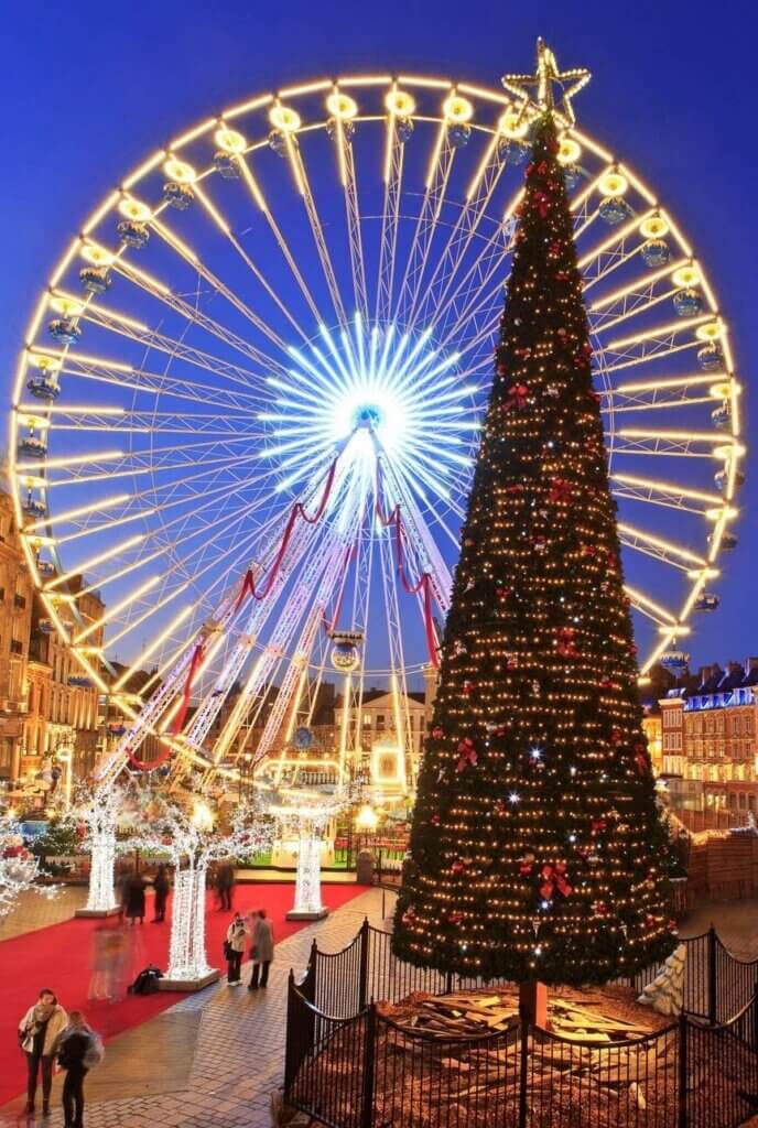 Twilight at Lille Christmas market, showcasing a radiant Ferris wheel and a large, ornately decorated Christmas tree, casting a warm glow over the festive stalls and happy visitors milling about the square.