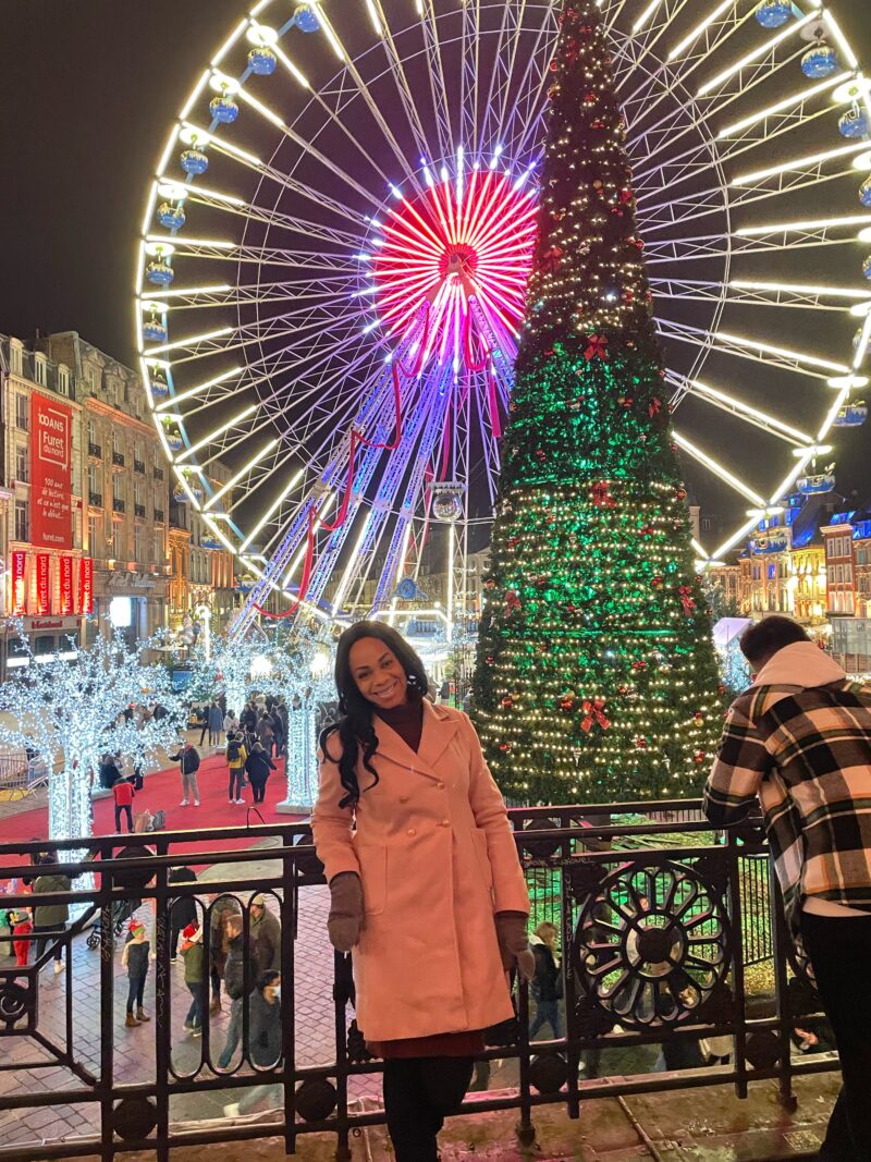 A smiling woman in a chic pink coat stands before the vibrant Lille Christmas market scene, with a spectacular Ferris wheel and a grandly decorated Christmas tree adding to the festive atmosphere.