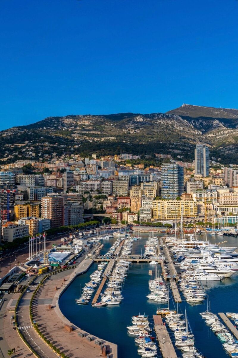 One Day in Monaco: A stunning vertical aerial view of the harbor in Monaco, displaying rows of moored yachts, a curved breakwater, and a densely populated cityscape climbing up the mountainous terrain under a clear blue sky.