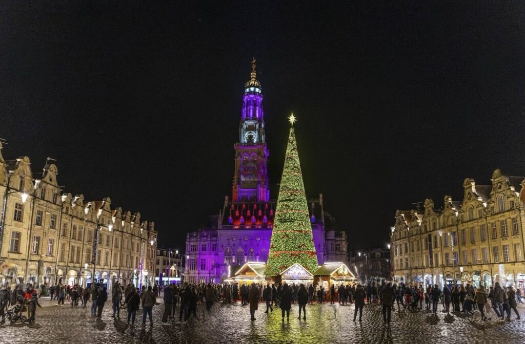 A bustling evening scene at one of the best French Christmas markets in Arras, with a magnificent Christmas tree and the belfry tower dramatically lit against the night sky, surrounded by the classical facades of town square buildings.