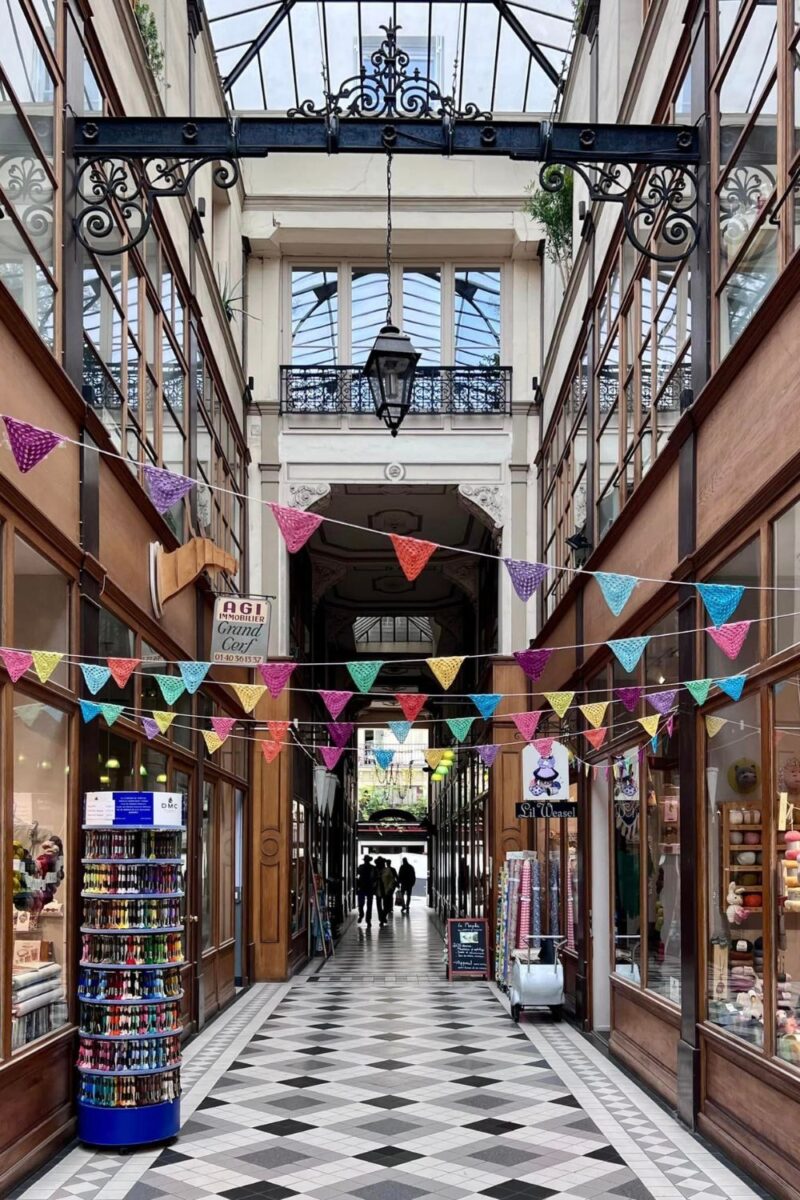 Interior view of a covered shopping arcade in Paris with black and white checkered flooring, colorful bunting flags strung overhead, and boutique shopfronts lining the corridor. Silhouettes of people are visible in the distance under a glass roof, highlighting the arcade's bustling atmosphere.