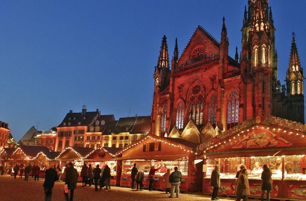 Twilight falls over one of the best French Christmas markets in Mulhouse, with a Gothic church bathed in red light providing a dramatic backdrop to the festive stalls and the soft glow of holiday lights.