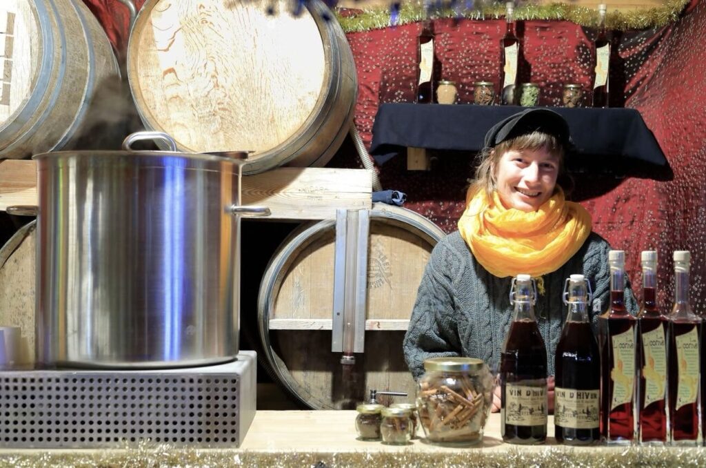 A smiling vendor in a yellow scarf and black hat serves mulled wine at the Reims Christmas Market, surrounded by wooden wine barrels and bottles of 'Vin d'Hiver' on display, exuding a warm and inviting holiday atmosphere.