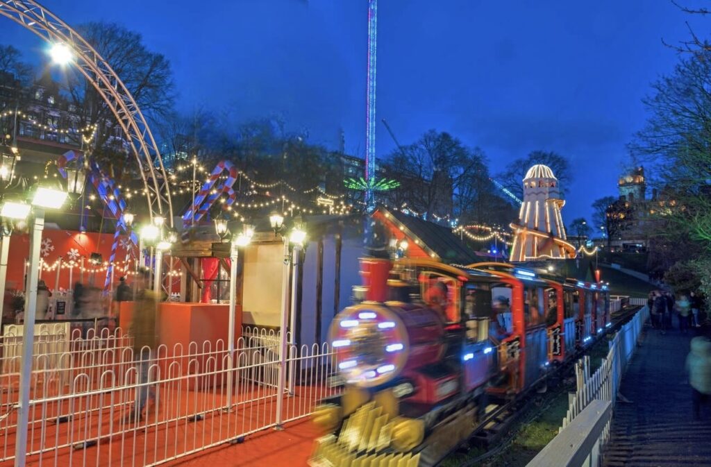 A festive children's train ride bustling with activity at the Reims Christmas Market during twilight, with colorful lights and decorations, an amusement ride, and an illuminated Ferris wheel in the background.