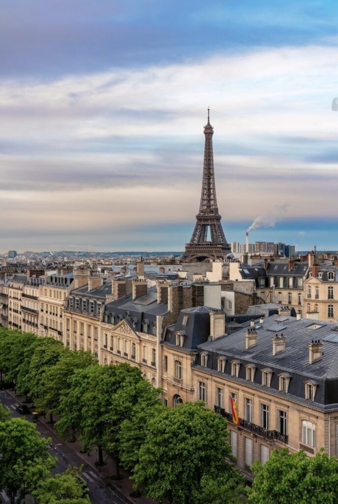 The iconic Eiffel Tower stands tall over the Parisian cityscape at dusk, with the warm glow of the sunset touching the historical Haussmannian buildings and the lush green trees lining the streets.