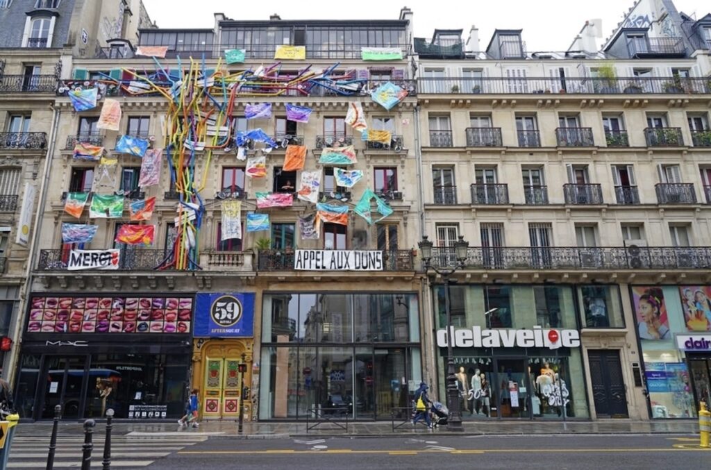 hidden gems in paris: Urban art installation at 59 Rivoli in Paris, featuring a colorful array of banners and ribbons streaming from a central point on the building's facade, with signs reading 'MERCI' and 'APPEL AUX DONS'. Below, the street-level shops display vibrant storefronts, including a MAC cosmetics store with a window display of lipstick shades.