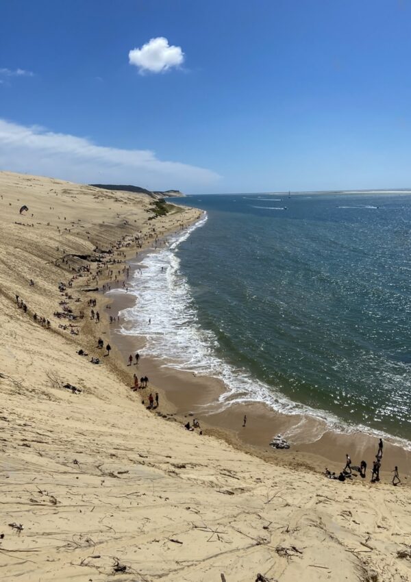 A striking view from the top of the Dune of Pilat in Gironde, France, showing visitors descending the sandy slope to the beach below, where the waves gently meet the shore. The dune's grand scale is juxtaposed with the vast blue sea and a single cloud in the bright sky above.