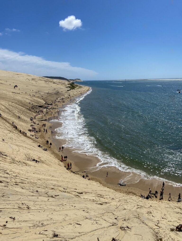 A striking view from the top of the Dune of Pilat in Gironde, France, showing visitors descending the sandy slope to the beach below, where the waves gently meet the shore. The dune's grand scale is juxtaposed with the vast blue sea and a single cloud in the bright sky above.