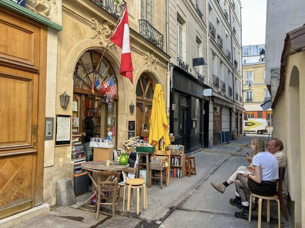 The outdoor seating area of La REcyclerie in Paris, bustling with people socializing at wooden tables next to an old railway track overgrown with greenery. The unique urban hangout spot showcases a creative use of space with a lush mini-garden alongside the repurposed tracks.