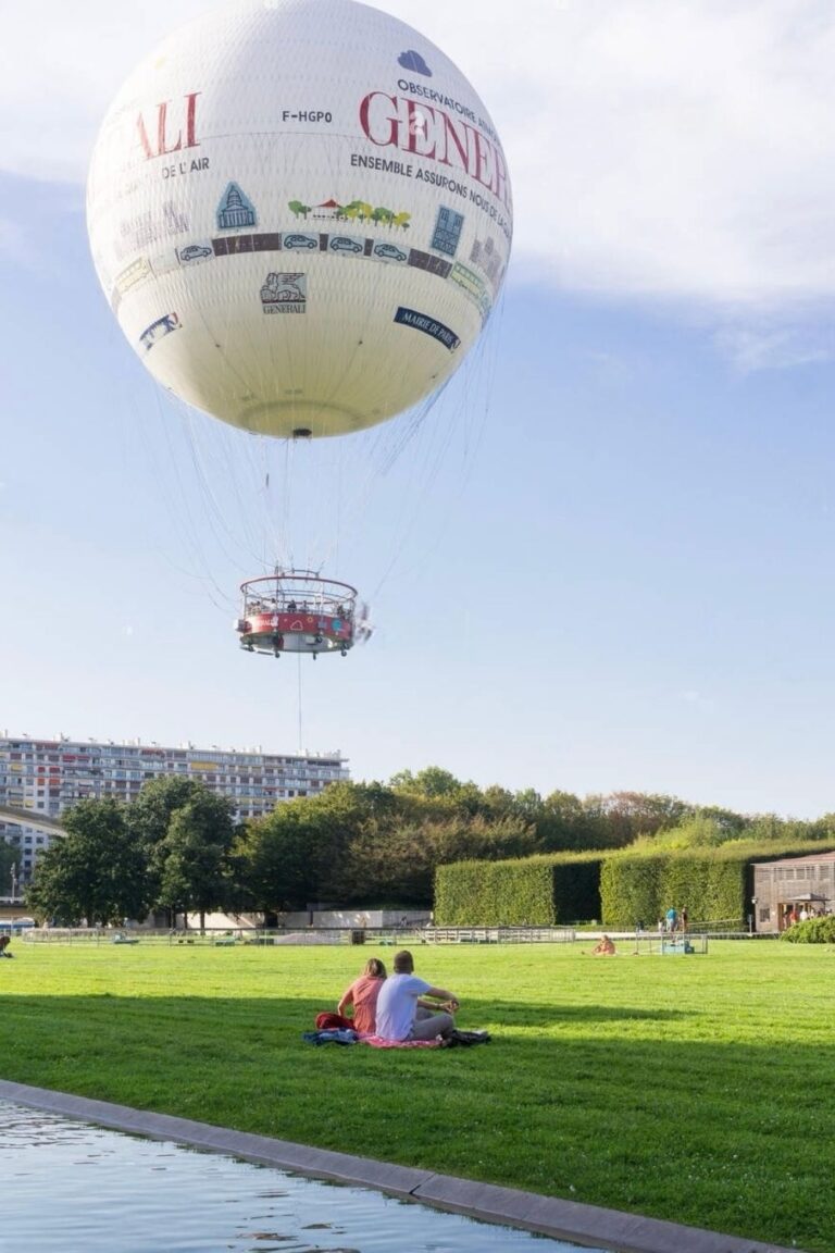 A couple sits on the grass in Parc André Citroën, enjoying a leisurely day with the 'Ballon de Paris Generali' tethered hot air balloon floating above. The park offers a tranquil setting for relaxation under the Paris sky, with the balloon providing a whimsical touch to the cityscape.