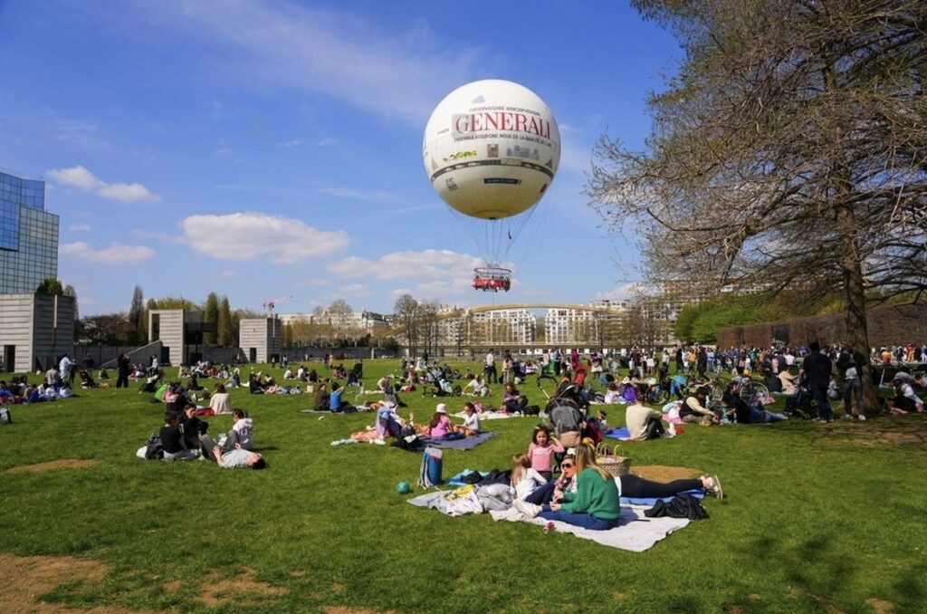 A vibrant day at the park with the 'Ballon de Paris Generali' hot air balloon floating above a relaxed crowd enjoying picnics on the grass. The clear blue sky and modern glass buildings in the background contrast with the leisurely atmosphere at ground level.