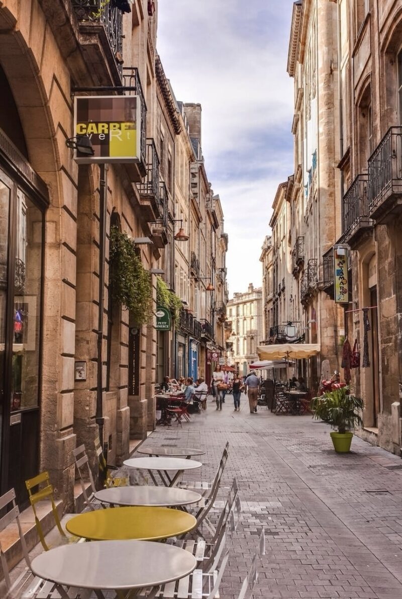 A quaint street in Bordeaux, lined with traditional limestone buildings and inviting café terraces. The picture captures the relaxed ambiance of city life, with people dining and strolling, and vibrant shop signs adding a pop of color to the historic charm of the pedestrian-friendly cobblestone path.