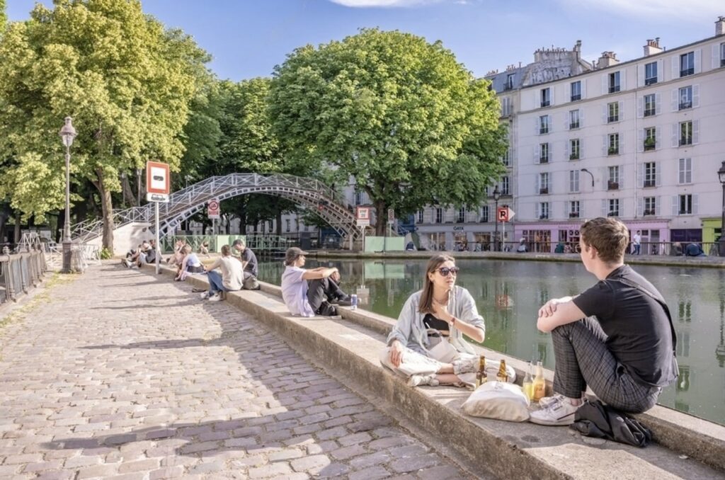 People relaxing along the Canal Saint-Martin in Paris, with some seated on the edge enjoying conversations and a picnic, overlooking the calm water. A classic Parisian iron footbridge crosses the canal in the background, framed by lush green trees and traditional apartment buildings.