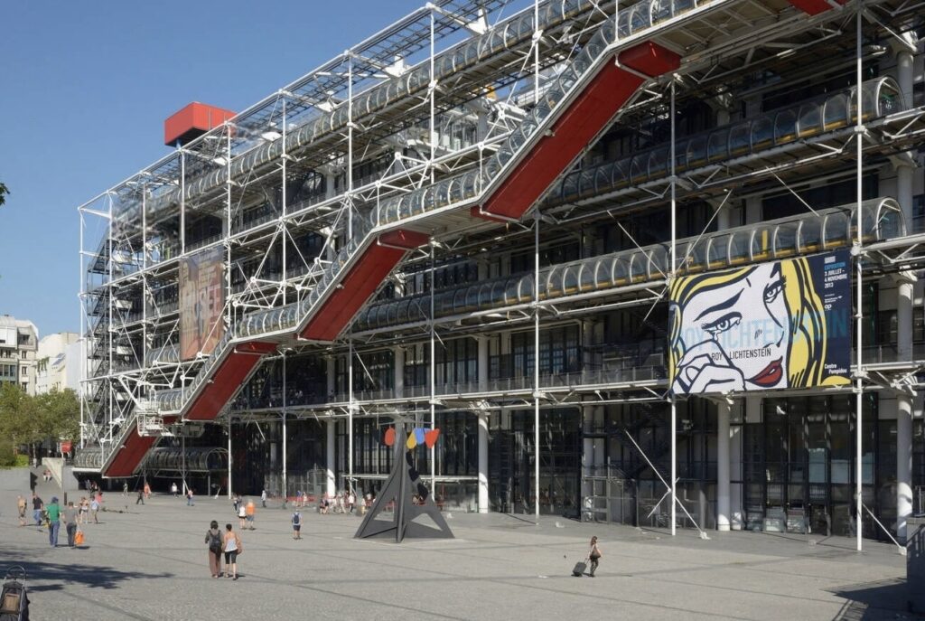 Exterior view of Centre Pompidou in Paris, showcasing its high-tech architecture with visible structural elements and colorful escalators, with people milling about in the foreground and a Roy Lichtenstein exhibition banner.