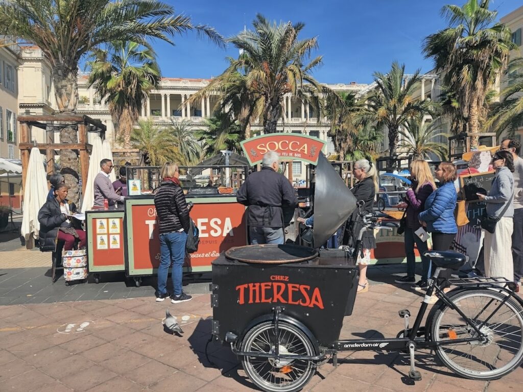 Bustling outdoor scene at Chez Thérèsa, a popular socca stand in Nice, France, with customers queuing for the traditional chickpea pancake, cooked over a wood fire. The backdrop features palm trees and classic Mediterranean architecture, capturing the lively street food culture.