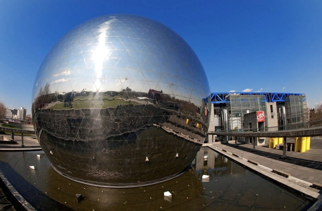 The Geode, a mirror-finished geodesic dome reflecting the surrounding park and architecture, with the clear blue sky overhead and the Cité des Sciences building in the background, Paris.